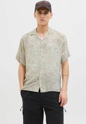 Chemise à manches courtes, de couleur vert clair, boutonnée, avec un motif semblable à de l'eau ; fabriquée dans un tissu léger et transparent, associée à un pantalon cargo noir.