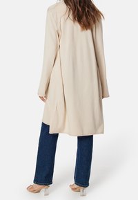 Beżowy długi kardigan z otwartym przodem, miękka struktura i długie rękawy, w połączeniu z ciemnoniebieskimi jeansami typu straight-leg i przejrzystymi sandałami na obcasie.