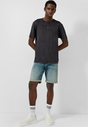Junger Mann steht mit den Händen hinter dem Rücken, trägt ein dunkles Henley-Shirt mit kurzen Ärmeln, hochgekrempelte Jeansshorts, weiße Socken und weiße Sneakers.