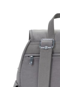 Gros plan sur une sangle de sac à dos gris avec une boucle en métal et un tirette de fermeture éclair sur une poche en tissu, sur fond blanc.
