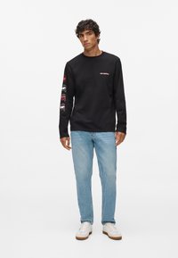 Jauns vīrietis stāv, ģērbts melnā garo piedurkņu krekla ar sarkaniem un baltajiem grafiskajiem ikonām uz piedurknēm, gaiši zilos džinsos un baltās sporta kurpēs.