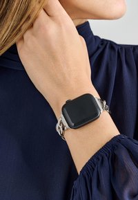 Zegarek smartwatch z kwadratową, czarną tarczą, srebrną metalową bransoletą z przeplatającymi się ogniwami, noszony na nadgarstku w zestawie z teksturowaną granatową bluzką.