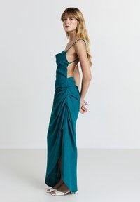 Frau in teal farbenem, gerafftem, rückenlosen Kleid mit Spaghettiträgern, steht seitlich mit gefalteten Händen hinten, trägt weiße Sandaletten mit offenem Zeh.
