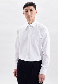 Wit overhemd met lange mouwen en een button-down kraag, voorzien van een borstzak en een ontspannen pasvorm, gecombineerd met donkere, geplooide broek.