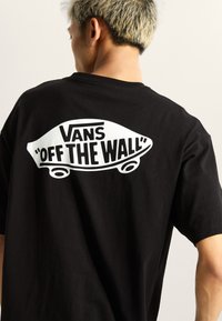 Camiseta de algodón negra con un gran gráfico blanco de una patineta y el texto "VANS 'OFF THE WALL'" en la parte posterior.