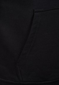 Sudadera negra con tejido que presenta un bolsillo destacado, textura suave, costuras limpias y puños acanalados en el dobladillo.