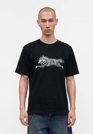 Mand iført en sort T-shirt med et sølvprikket motiv af en løbende tegneserieulv på fronten, kombineret med blå jeans og en kædeaccessory.