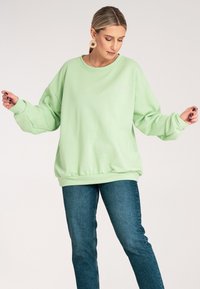 Jasnozielony, oversize'owy sweter z okrągłym dekoltem, ściągaczami przy mankietach i u dołu. Z przodu ma mały różowy napis. W zestawie z ciemnoniebieskimi dżinsami.