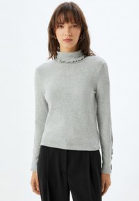 Grijze coltrui met ruches aan de hals, lange mouwen en zachte structuur. Gekozen in combinatie met zwarte broek, wat een casual uitstraling geeft.
