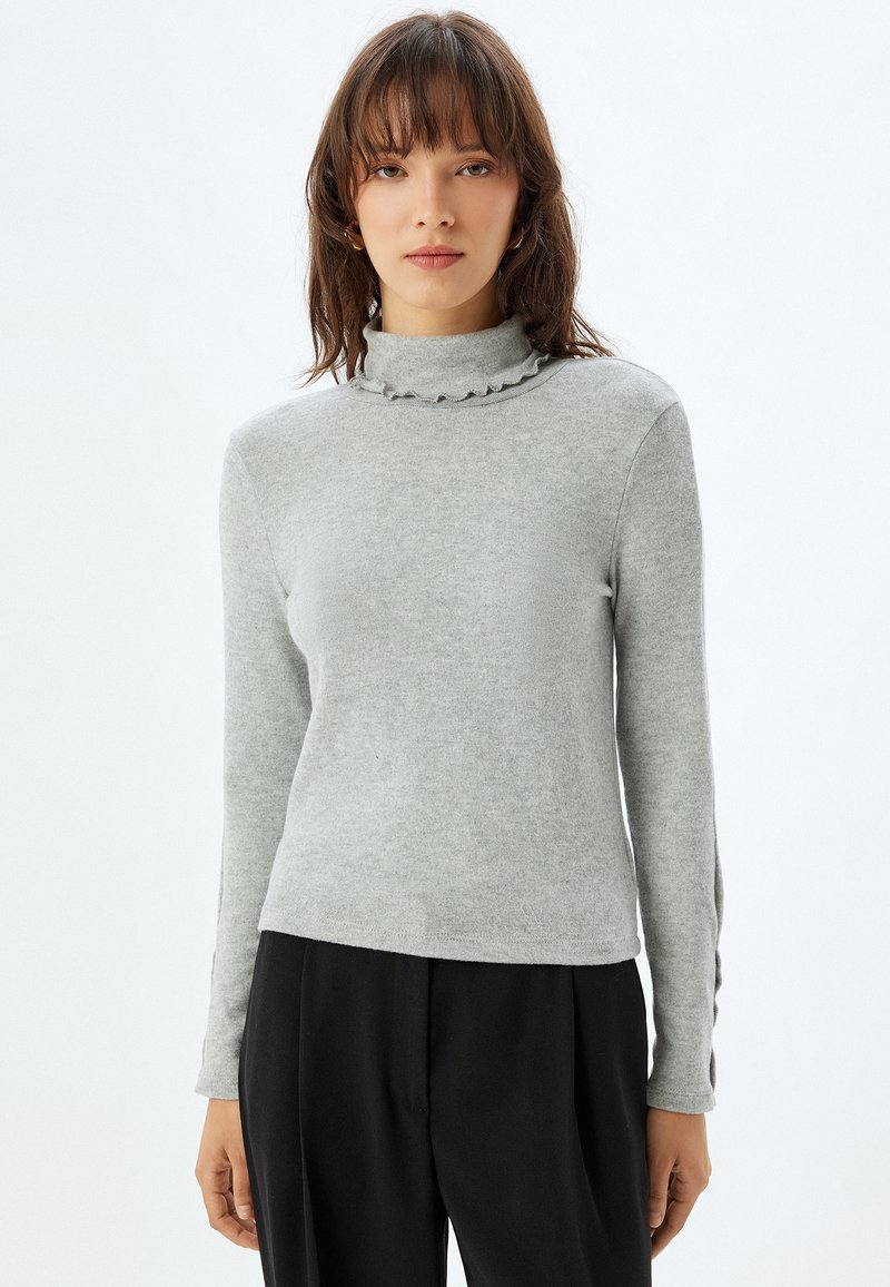 Grijze coltrui met ruches aan de hals, lange mouwen en zachte structuur. Gekozen in combinatie met zwarte broek, wat een casual uitstraling geeft.