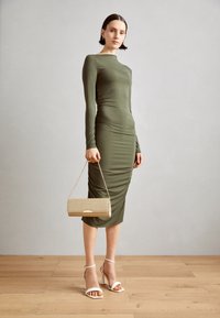 Olivově zelené dlouhé šaty s řasením, s kombinací zlaté třpytivé kabelky a bílých sandálů na podpatcích na dřevěné podlaze.