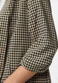 Tkanina s kaširanom hounds-tooth uzorkom u bež i crnoj boji, s naboranom šavom na rukavu do tri četvrtine, djelomično pokriva ruku.