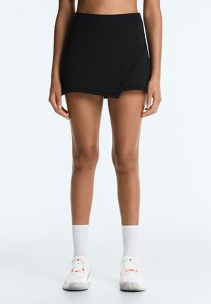 Žena v čiernom asymetrickom skorte, bielych členkových ponožkách a bielych teniskách, stojaca s uvoľnenými rukami vedľa tela na jednoduchom pozadí.