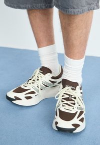 Baskets de sport blanches et marron avec une tige en mesh, semelle extérieure noire contrastante et des motifs texturés. Portées avec des chaussettes blanches.