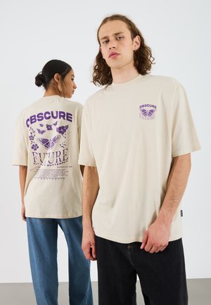 Beige oversized t-shirts med lilla grafiske designs, der viser sommerfugle og blomster. Modellen til venstre har blå jeans, mens modellen til højre har sorte jeans.