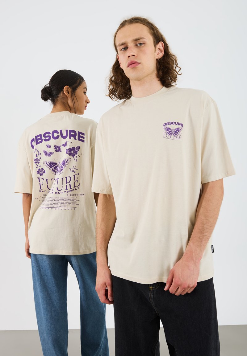 Beige oversized t-skjorter med lilla grafiske design som viser sommerfugler og blomster. Modellen til venstre har på seg blå jeans, mens modellen til høyre har på seg svarte jeans.