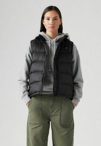 Chaleco acolchado negro con un cuello alto, cierre de cremallera y bolsillos laterales, llevado sobre una sudadera gris, acompañado de pantalones verde oliva.