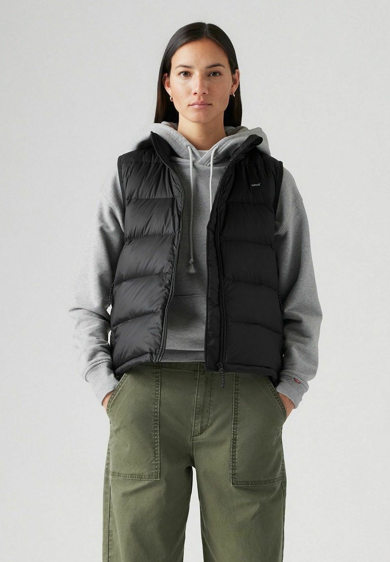 Chaleco acolchado negro con un cuello alto, cierre de cremallera y bolsillos laterales, llevado sobre una sudadera gris, acompañado de pantalones verde oliva.