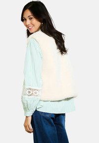 Crème fleece vest met een ronde halslijn, gecombineerd met een lichtgroen en wit gestreept shirt met kantaccenten. Casual blauwe jeans gedragen.