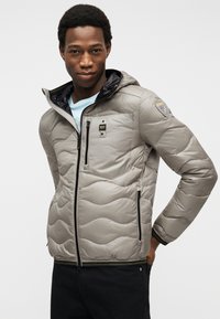 Νέος άνδρας που φοράει ανοιχτό γκρι μπουφάν με κουκούλα και μαύρες τσέπες με φερμουάρ, στέκεται μπροστά σε απλό λευκό φόντο.