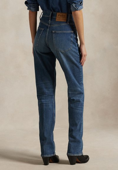 Asmuo dėvi tamsiai mėlynas džinsines kelnės aukštu liemeniu su užpakalinėmis kišenėmis ir juodus aukštakulnius batus, stovintis prieš paprastą foną.