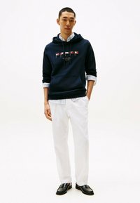 Granatowy hoodie z kieszenią z przodu, ozdobiony kolorowym logo. W zestawieniu z białymi spodniami i czarnymi mokasynami, podkreślającymi swobodny styl.