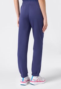 Pantalones de chándal morados con cintura elástica y puños ajustados, hechos de tela suave. Llevados con zapatillas blancas con acentos en rosa y azul.