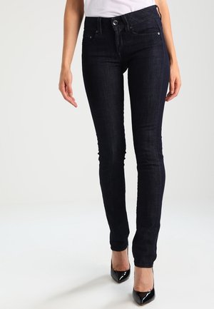 Vrouw draagt donkerblauwe skinny jeans, witte top en zwarte spitse hoge hakken, staat met één voet iets naar voren.