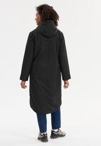 Manteau noir long avec capuche, présentant une texture lisse et des poches latérales. Associé à un jean en denim bleu et des baskets grises.