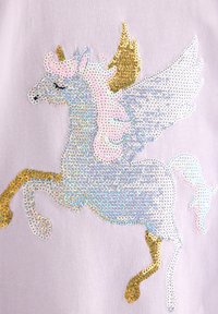 Μονόκερος με χρυσή τρίχα και οπλές, ροζ χαίτη και λευκά φτερά από γυαλιστερές παγιέτες σε ανοιχτό μωβ ύφασμα.