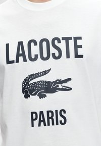 Chemise blanche avec texte noir "LACOSTE" en haut et texte "PARIS" en dessous d'un graphisme détaillé de crocodile noir.