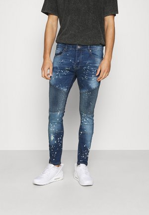 Homme portant un jean slim bleu avec des panneaux côtelés au niveau des genoux et un motif éclaboussé de peinture blanche, assorti à des baskets blanches et une chemise sombre.