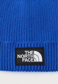 Πλεκτός σκουφί σε ζωηρό μπλε με ριμπ υφή. Διαθέτει ένα εμφανές μαύρο ετικέτα με το λογότυπο "THE NORTH FACE" σε λευκό.