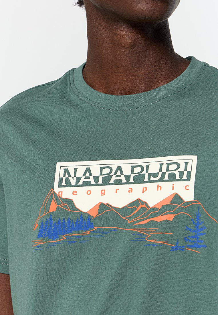 Man in een groen T-shirt met het "NAPAPIJRI geographic"-logo en een grafisch ontwerp van oranje bergen en blauwe bomen op de voorkant.
