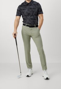 Černá vzorovaná polo košile spárovaná se světle zelenými golfovými kalhotami. Oblečení doplňuje černý opasek a bílé golfové boty. Hůl v ruce.