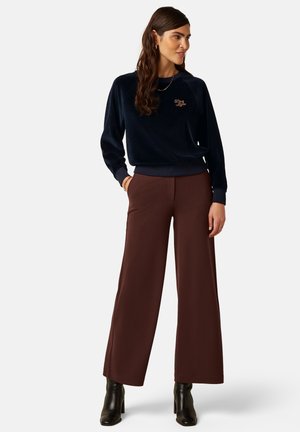 Kvinde iført en navyblå sweatshirt med tekst og brune vidde bukser, står med den ene hånd i lommen og sorte ankelstøvler mod hvid baggrund.