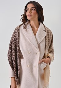 Cappotto di lana doppiopetto color crema con tasche frontali, indossato con uno scialle a motivo marrone e beige con frange lungo i bordi.