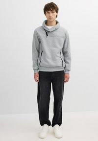 Sudadera gris con un alto cuello y detalles con cremallera, combinada con jeans de mezclilla negros y zapatillas blancas. Textura suave, ajuste casual.