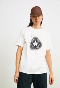 Camiseta de algodón blanca con un gráfico circular negro que presenta una estrella y el texto "Converse All Star". Se lleva con una gorra marrón. Se pueden ver pantalones oscuros.