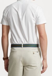 Man som bär en kortärmad vit skjorta, beige byxor med bakfickor, randigt bälte och brun klocka, sedd bakifrån.