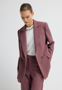Op maat gemaakt tweedelig pak van mauve stof. Opvallende kenmerken zijn een enkelzijdig blazer, twee voorzakken en vergulde hardware accenten.