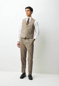 Beige ternet tre-delt kostume med en tætsiddende vest, knaplukning og matchende bukser, parret med en hvid skjorte og brunt slips. Sorte sko.