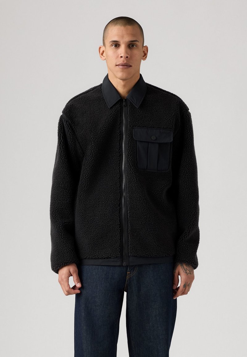Zwarte fleece jas met een ritssluiting, een zwarte stoffen kraag en een borstzak. Kenmerkt zich door een gestructureerde stof en een casual pasvorm. Gedragen over denim.