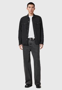 Schwarzes Button-Up-Hemd mit zwei Brusttaschen, getragen über einem weißen T-Shirt, kombiniert mit grauen weit geschnittenen Jeans und schwarzen Schuhen. Schlichtes Design.