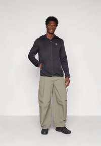 Homem a usar um hoodie preto com fecho, calças largas bege e sapatos pretos, de pé com uma mão no bolso contra um fundo simples.