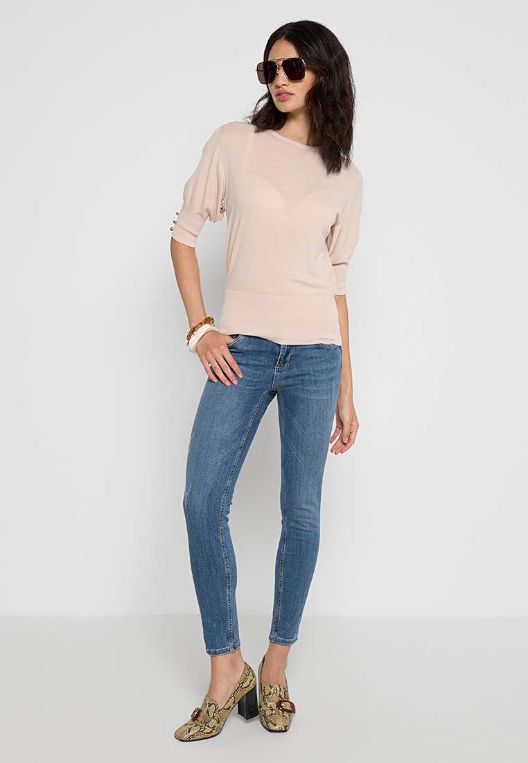 Heller rosa gestrickter Sweater mit kurzen Ärmeln, kombiniert mit eng anliegenden blauen Jeans. Das Modell trägt gemusterte braune und goldene Blockabsatzschuhe.