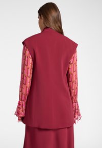 Blazer senza maniche color bordeaux con spalle enfatizzate, sovrapposto a maniche rosa a fantasia con polsini arricciati. Tessuto dalla texture liscia.