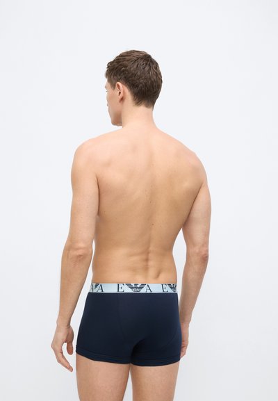 Νέος άνδρας στέκεται πλάτη στην κάμερα, φοράει μποξεράκια σε σκούρο μπλε χρώμα με ανοιχτόχρωμη μέση που έχει λογότυπα.