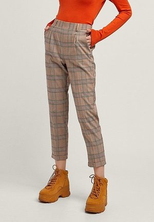 Vrouw met hoge taille bruine geruite broek, oranje top met lange mouwen en mosterdgele platform veterlaarzen, handen in de zakken.