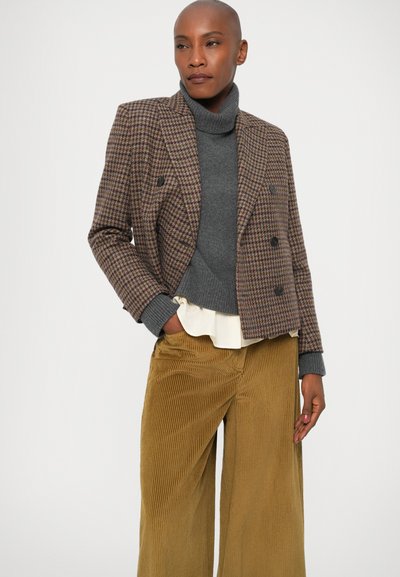 Blazer a cuadros marrón con botones de doble botonadura, cuello de tortuga gris, blusa blanca en capas y pantalones de pana de pierna ancha en color mostaza.